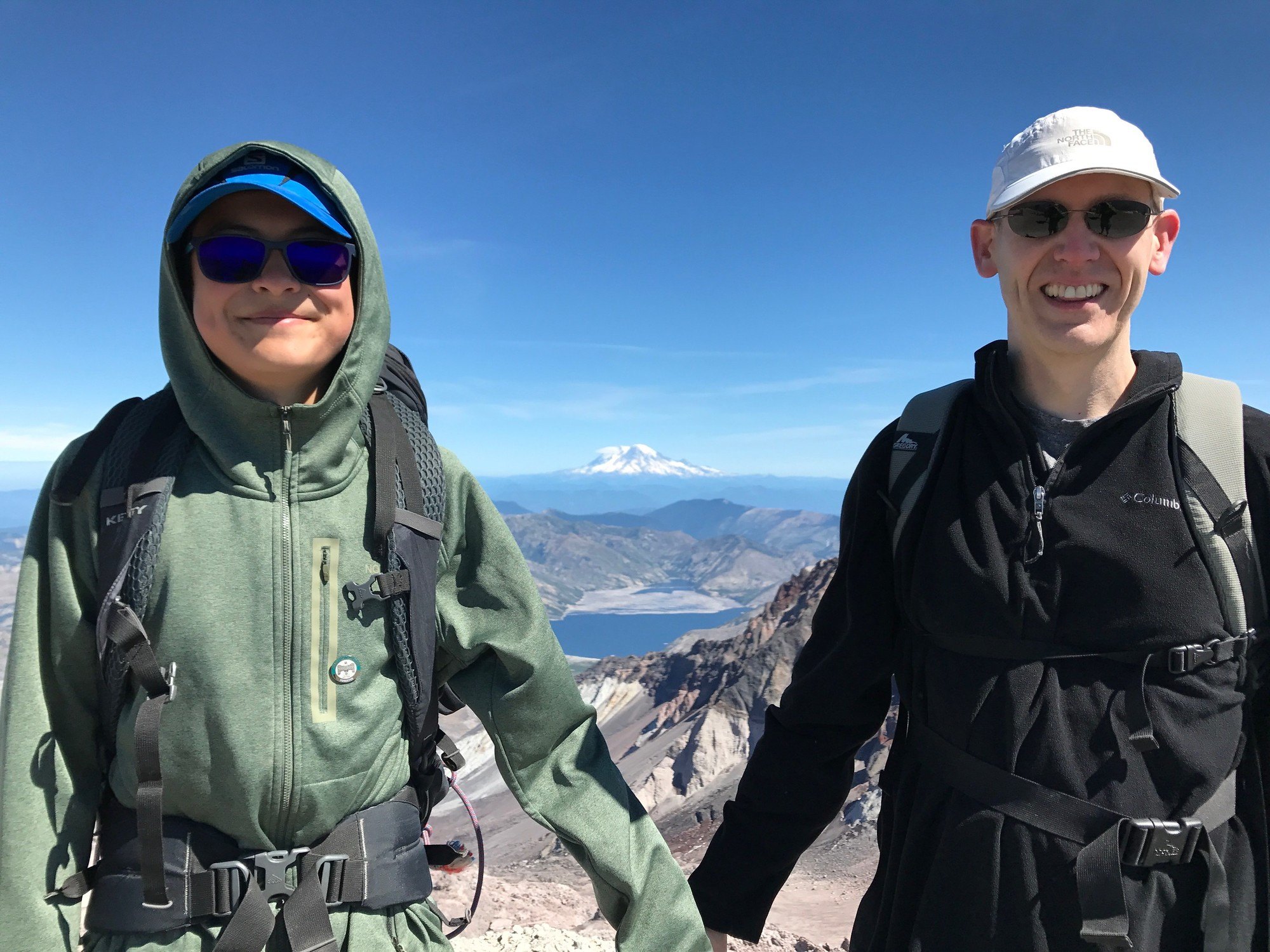 Mark Nelson and teenage son smiling on a rocky mountain top with a lake and Mount Rainier in the background
