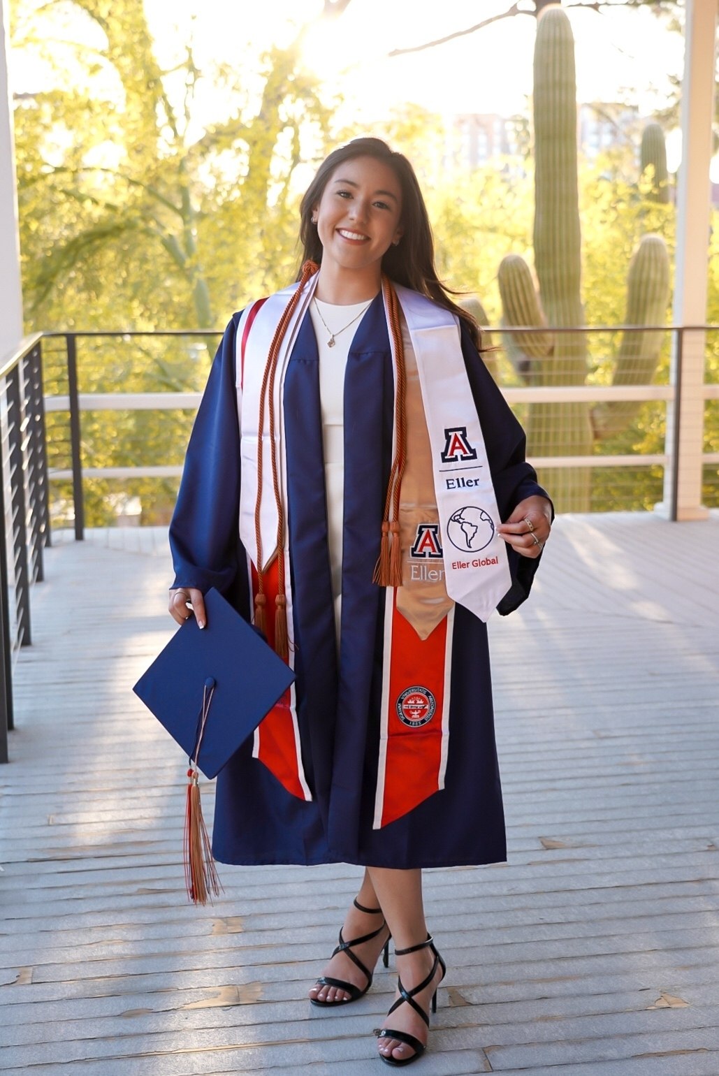 Smiling young woman wearing blue graduation gown, holding cap in one hand and Eller College of Management stole in the other