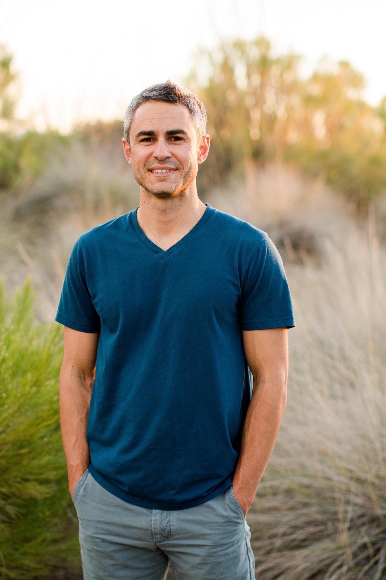 Man in blue t-shirt, sage green pants, short hair ,standing in a grassy field.