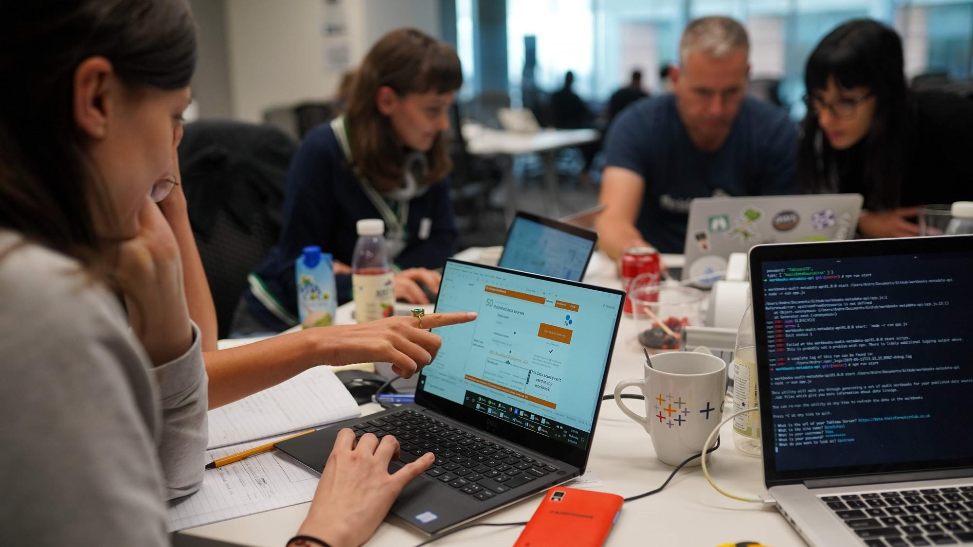 Image of three women and one man sitting at a table using Tableau and other coding softwares. 