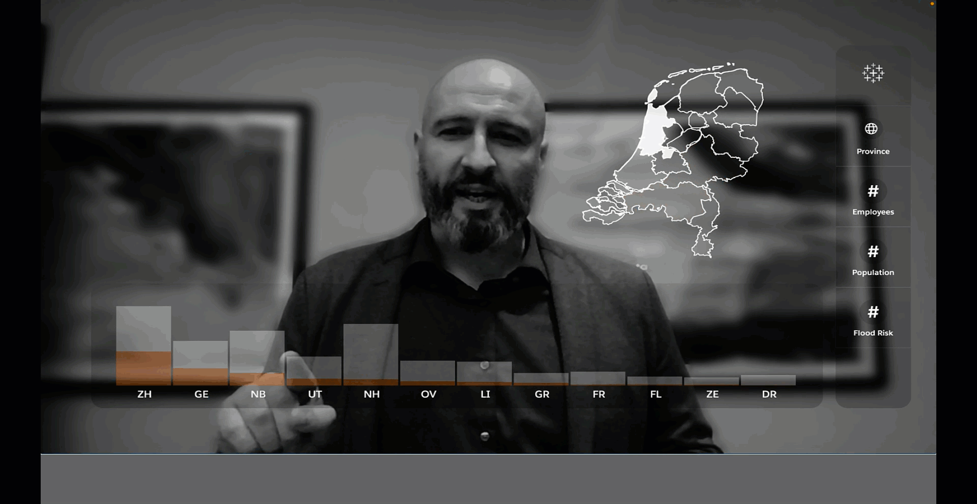 Black and white image of man in dark, buttoned shirt and blazer, pointing at bar charts and map on a screen