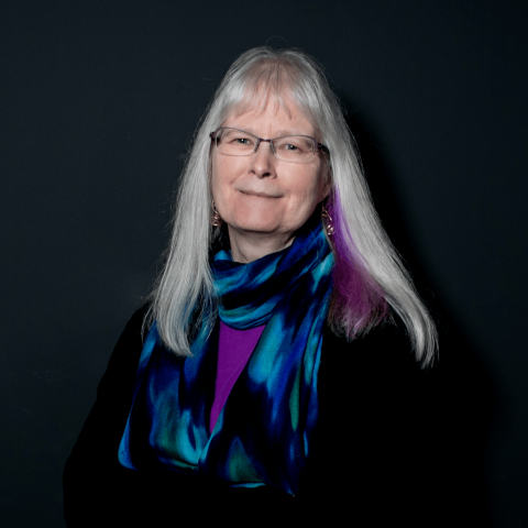 Headshot of a woman with grey hair and a purple dye streak wearing a black sweater with a colorful scarf