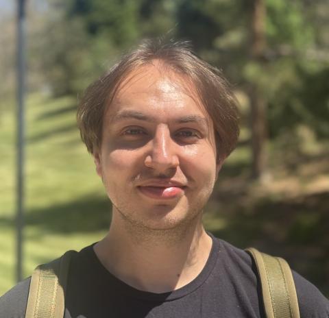Portrait photo of Zach Cutler outdoors in sunlight, smiling and wearing a black shirt with backpack straps visible, against a softly blurred background of trees and grass.