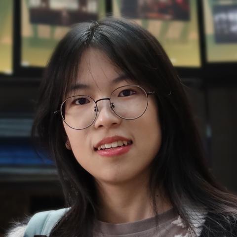 Portrait photo of Manling Yang smiling indoors, wearing round glasses and a light-colored top, with a softly blurred background featuring framed displays.
